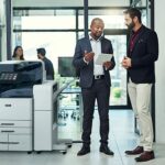 Two men in an office setting discussing something on a tablet beside a Xerox® AltaLink® C8200 Series Colour Multifunction Printer