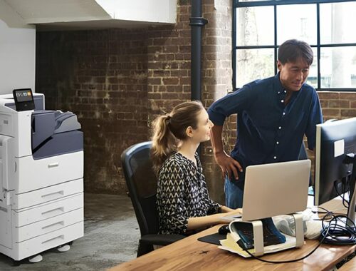 A man and woman stand side by side in front of a computer, focused on the screen. The Xerox® VersaLink® C7000 Colour Printer is on the background.